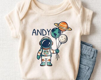 May include: A cream-colored short-sleeve bodysuit featuring a graphic of an astronaut holding three balloons shaped like Earth, the moon, and Saturn. The name ANDY is printed in dark blue letters above the astronaut figure.