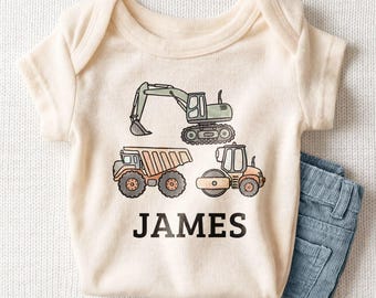 May include: A cream-colored short-sleeve bodysuit featuring a graphic print of a green excavator, an orange dump truck, and an orange steamroller. The name JAMES is printed in bold black capital letters below the construction vehicles.
