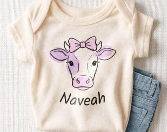 May include: A cream-colored short-sleeve bodysuit featuring a line-art illustration of a cow with a pink bow and purple accents. The name Naveah is printed in black script lettering below the cow graphic, paired with blue corduroy pants.