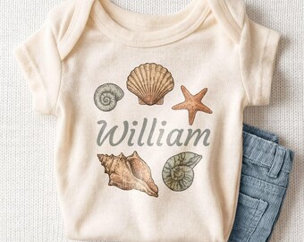 May include: A cream-colored short-sleeve bodysuit featuring a graphic print of various seashells and a starfish surrounding the name William in a cursive font, paired with a pair of blue corduroy pants.