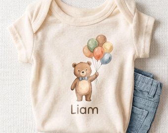 May include: A cream-colored short-sleeve baby bodysuit featuring a brown bear illustration wearing a blue bow tie and holding a cluster of five multicolored balloons. The name Liam is printed in brown text below the bear. A pair of blue corduroy pants.