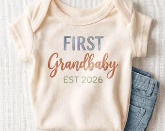 May include: A cream-colored short-sleeve baby bodysuit featuring the text FIRST in blue, Grandbaby in a brown cursive script, and EST 2026 in green. The garment is paired with a pair of blue corduroy pants visible at the bottom right corner.