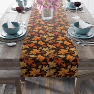 May include: A table runner with a repeating pattern of autumn leaves in shades of orange, yellow, and brown on a dark background. The runner is placed on a wooden table set for a meal, with plates, glasses, and silverware.