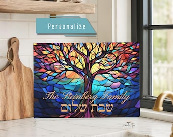 May include: A rectangular glass cutting board featuring a vibrant stained-glass style tree design with multicolored leaves in shades of blue, red, yellow, and purple. The text "The Steinberg Family" and Hebrew text "שבת שלום" are printed in gold.
