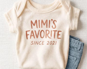 May include: A cream-colored short-sleeve baby bodysuit featuring the text MIMI'S FAVORITE SINCE 2021 in a copper-colored font, paired with a pair of blue corduroy pants with visible vertical texture and a button detail.
