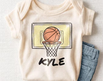 May include: A cream-colored short-sleeve baby bodysuit featuring a graphic print of a basketball above a hoop and backboard. The name KYLE is printed in bold black capital letters below the basketball hoop illustration.