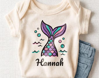 May include: A cream-colored short-sleeve bodysuit featuring a graphic of a purple, pink, and teal mermaid tail with bubbles and waves. The name Hannah is printed in black script lettering below the tail. A pair of blue corduroy pants is included.
