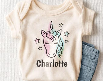 May include: A cream-colored short-sleeve baby bodysuit featuring a black line-art illustration of a unicorn head with soft pink and blue watercolor accents, surrounded by small stars, with the name Charlotte printed in black below.