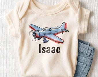 May include: A cream-colored short-sleeve baby bodysuit featuring a blue airplane illustration with red accents and the name Isaac printed in bold black letters below the graphic, paired with blue corduroy pants.