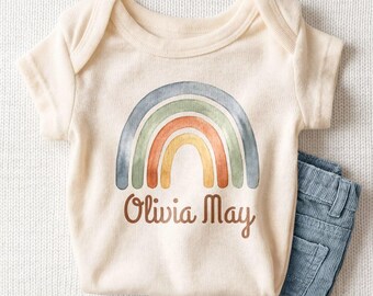 May include: A cream-colored short-sleeve bodysuit featuring a watercolor-style rainbow graphic in muted tones of blue, green, orange, and yellow. Below the rainbow, the name Olivia May is printed in a brown, cursive script font.