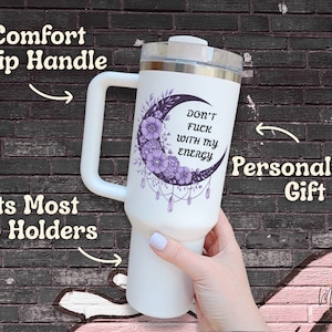 May include: White tumbler with a silver lid and a large white handle. The tumbler features a purple crescent moon design with flowers and the text "DON'T FUCK WITH MY ENERGY". The tumbler is a personalized gift.