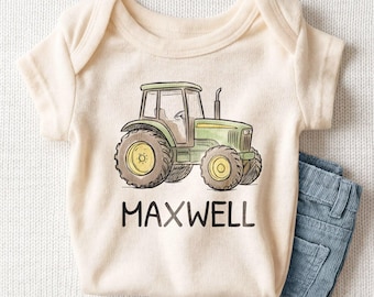 May include: A cream-colored short-sleeve bodysuit featuring a green and yellow tractor illustration centered on the chest. Below the tractor, the name MAXWELL is printed in bold, black, uppercase sans-serif capital letters.
