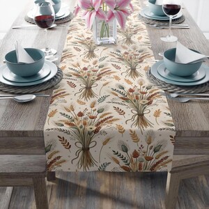 May include: A beige table runner with a repeating pattern of wheat and floral bouquets tied with ribbons. The runner is set on a wooden table with place settings, including blue bowls, plates, and wine glasses. A vase of pink lilies is in the center.