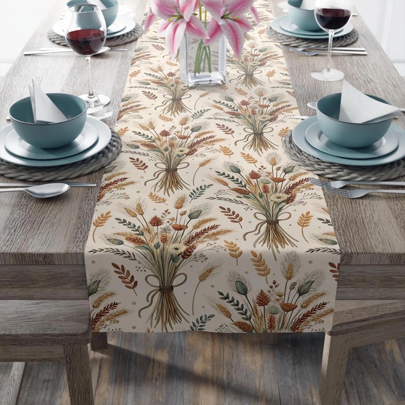 May include: A beige table runner with a repeating pattern of wheat and floral bouquets tied with ribbons. The runner is set on a wooden table with place settings, including blue bowls, plates, and wine glasses. A vase of pink lilies is in the center.