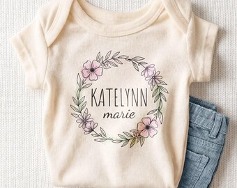 May include: A cream-colored short-sleeve baby bodysuit featuring a floral wreath graphic in black, pink, and green. The text KATELYNN marie is printed in the center of the wreath. A pair of blue corduroy pants is folded next to the bodysuit.