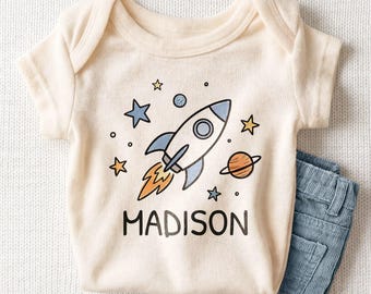 May include: A cream-colored short-sleeve baby bodysuit featuring a graphic of a rocket ship, planets, and stars in blue, orange, and yellow. The name MADISON is printed in black capital letters below the rocket. A pair of blue corduroy pants.