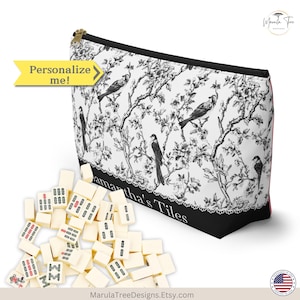 May include: A white and black zippered pouch with a bird and floral design, next to a pile of ivory-colored tiles. The pouch has the text "Samantha's Tiles" and a yellow arrow that says "Personalize me!".