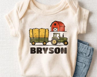May include: A cream-colored short-sleeve baby bodysuit featuring a graphic of a green tractor pulling a trailer loaded with yellow hay bales, a red barn, and the name BRYSON printed in bold black capital letters below the illustration.