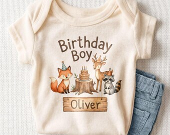 May include: A cream-colored short-sleeve bodysuit featuring a graphic of a fox, a deer, and a raccoon wearing party hats around a birthday cake on a tree stump. The text reads Birthday Boy and the name Oliver on a wooden sign.