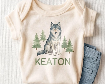 May include: A cream-colored short-sleeve bodysuit featuring a watercolor illustration of a gray wolf sitting among green pine trees. The name KEATON is printed in green capital letters below the wolf. A pair of blue corduroy pants is included.