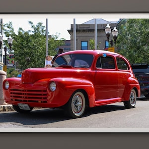 May include: A framed photograph of a classic red car parked on a city street. The vintage vehicle has a glossy finish, white-walled tires, and chrome accents. The background includes buildings, trees, and streetlights.