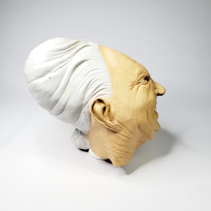 Puede incluir: Máscara de látex que muestra el perfil de una persona mayor con una expresión severa. La máscara presenta cabello blanco en el lado izquierdo y una cara bronceada con arrugas y la boca abierta. La máscara es probablemente para Halloween o disfraces.