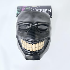 Puede incluir: Una máscara de plástico negra con una amplia sonrisa de dientes. La máscara tiene un diseño texturizado, similar a la fibra de carbono, y agujeros para los ojos cubiertos con una malla negra.