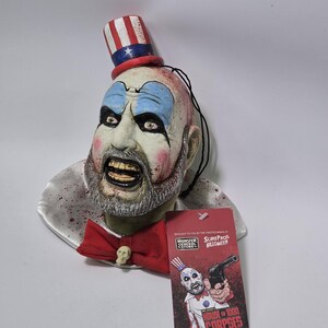 Può includere: Un busto da parete del Capitano Spaulding di House of 1000 Corpses, con volto pallido, trucco blu sugli occhi, barba grigia, cilindro rosso, bianco e blu, papillon rosso con teschio e colletto bianco con schizzi di sangue rosso.