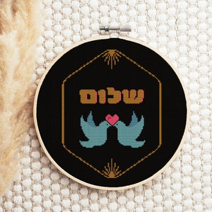 May include: A black embroidery hoop with a gold hexagonal frame. The frame features a stylized sunburst design at the top and bottom. Inside the frame, two blue birds are kissing with a red heart. The Hebrew word "Shalom" is written in gold letters above the birds.