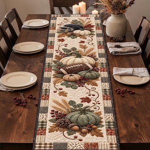 May include: A rectangular quilted table runner featuring a seasonal autumn-themed pattern with pumpkins, footballs, wheat stalks, pinecones, and leaves in shades of orange, brown, and green, bordered by a patchwork design of plaid and floral fabric squares.