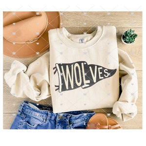 May include: A cream-colored sweatshirt with a black pennant design that says "WOLVES" in white letters. The sweatshirt is folded and laid on a wooden surface with a brown hat, blue jeans, and a small potted plant.