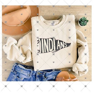 Könnte beinhalten: Ein cremefarbener Pullover mit dem Wort "INDIANS" in schwarzer Wimpelform darauf gedruckt. Der Pullover ist gefaltet und auf einer Holzoberfläche mit einer blauen Jeans, einem braunen Hut und einer kleinen Topfpflanze abgelegt.