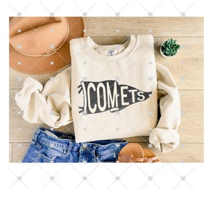 May include: A cream-colored sweatshirt with a black graphic of a pennant that says "COMETS". The sweatshirt is folded and laid on a wooden surface with a brown hat, blue jeans, and a small potted plant.