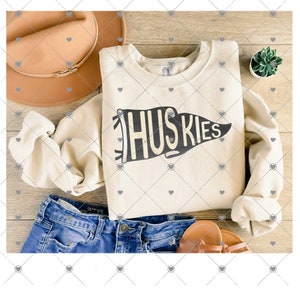 May include: A cream-colored sweatshirt with a black pennant design that says "HUSKIES". The sweatshirt is folded and laid out on a wooden surface with a brown hat, blue jeans, and a small potted plant.