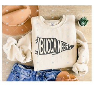 May include: A cream-colored sweatshirt with a black pennant design that says "BUCCANEERS". The sweatshirt is laid out on a wooden surface with a brown hat, blue jeans, and a small potted plant.