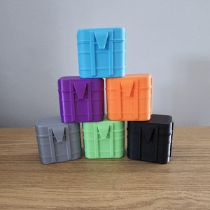 May include: Six small, stackable boxes in different colours: light blue, purple, orange, grey, green, and black. Each box has a design resembling a crate with a handle on the top.