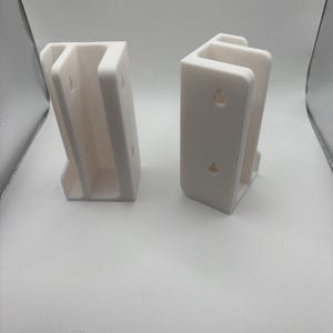 May include: Two white, U-shaped brackets with mounting holes. The brackets are made of a solid, opaque material and are designed to hold an object. The brackets are shown against a white background.