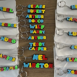 May include: A collection of colourful, personalised keychains with names spelled out in block letters. The keychains are made of a soft, flexible material and are attached to silver key rings.