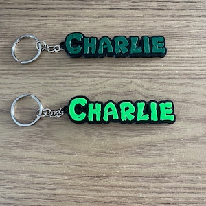 May include: Two keyrings, each featuring the name "Charlie" in bright green letters with a black outline. The keyrings have a silver chain and ring. They are displayed on a light brown wooden surface.