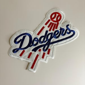 May include: Embroidered patch featuring the Los Angeles Dodgers logo. The patch has a white background with the word "Dodgers" in blue script. Red lines and a baseball graphic are above the word, and the patch is outlined in white.