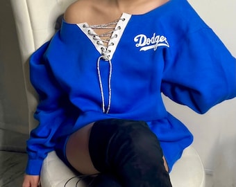 May include: A royal blue sweatshirt with a white lace-up detail at the neckline and the word "Dodger" in white lettering. The sweatshirt is worn with black thigh-high boots.