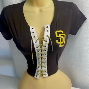 Pode incluir: Camiseta preta de manga curta com um detalhe de renda branca na frente. A camiseta tem um logotipo "SD" amarelo do lado direito.