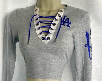May include: Gray long-sleeve crop top with a white lace-up neckline and gold eyelets. The top features a blue glitter "LA" logo on the front and a blue glitter "LA" logo on the sleeve.