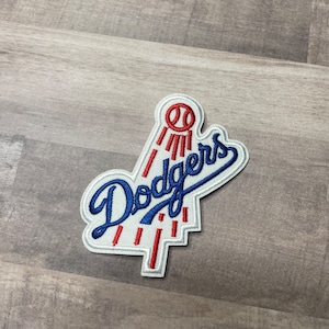 May include: Embroidered patch featuring the word "Dodgers" in blue script with a white outline. The patch includes a baseball and red accents, with a white background. The patch is approximately 3 inches wide.