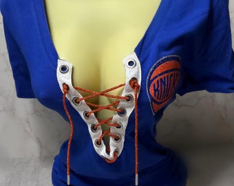 Puede incluir: Una camiseta azul con un logotipo de baloncesto blanco y naranja y un escote con cordones. Los cordones son rojos y blancos.