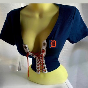 Puede incluir: Una camiseta azul marino de manga corta con un detalle de encaje blanco y rojo en la parte delantera. La camiseta tiene un gran logotipo "D" en el lado derecho.