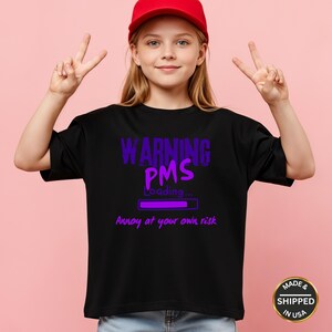 Könnte beinhalten: Schwarzes T-Shirt mit dem Text "WARNING PMS Loading... Annoy at your own risk" in Lila. Die Person auf dem Bild trägt eine rote Baseballkappe und zeigt mit beiden Händen das Friedenszeichen.