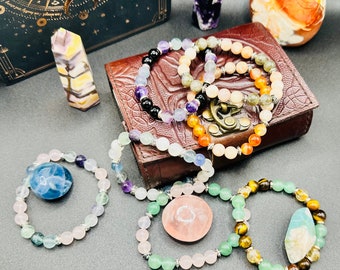 Op de afbeelding: Een verzameling kleurrijke edelsteen armbanden met verschillende kristallen en kralen. De armbanden zijn gerangschikt op een bruin lederen dagboek met een gouden ontwerp. Er zijn ook verschillende kristallen en een klein boek op de achtergrond.