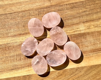 Op de afbeelding: Een verzameling van acht gladde, ovale rozenkwarts stenen. De stenen hebben een zachte roze kleur met subtiele variaties in toon en textuur. Ze liggen op een houten oppervlak, met natuurlijk licht dat schaduwen werpt.