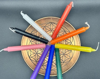 Op de afbeelding: Een verzameling van negen dunne, cilindrische kaarsen in diverse kleuren, waaronder rood, oranje, geel, groen, blauw, paars, roze, zwart en wit, gerangschikt in een radiaal patroon op een ronde, koperkleurige metalen schaal.